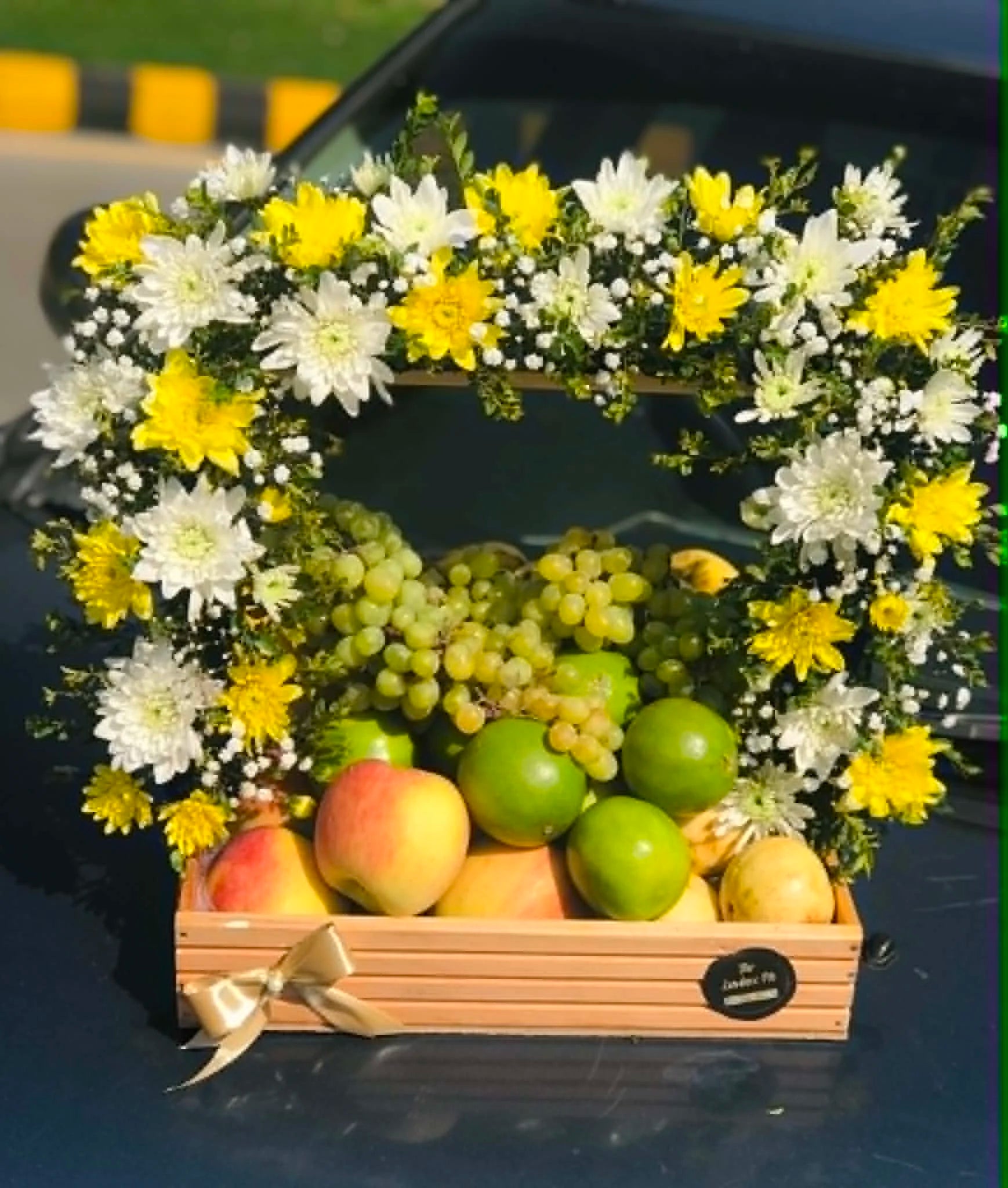 Luxury seasonal fruit basket with 3–5 kg mixed fruits, white and yellow flowers, and a premium wooden handle basket for gift delivery in Pakistan.