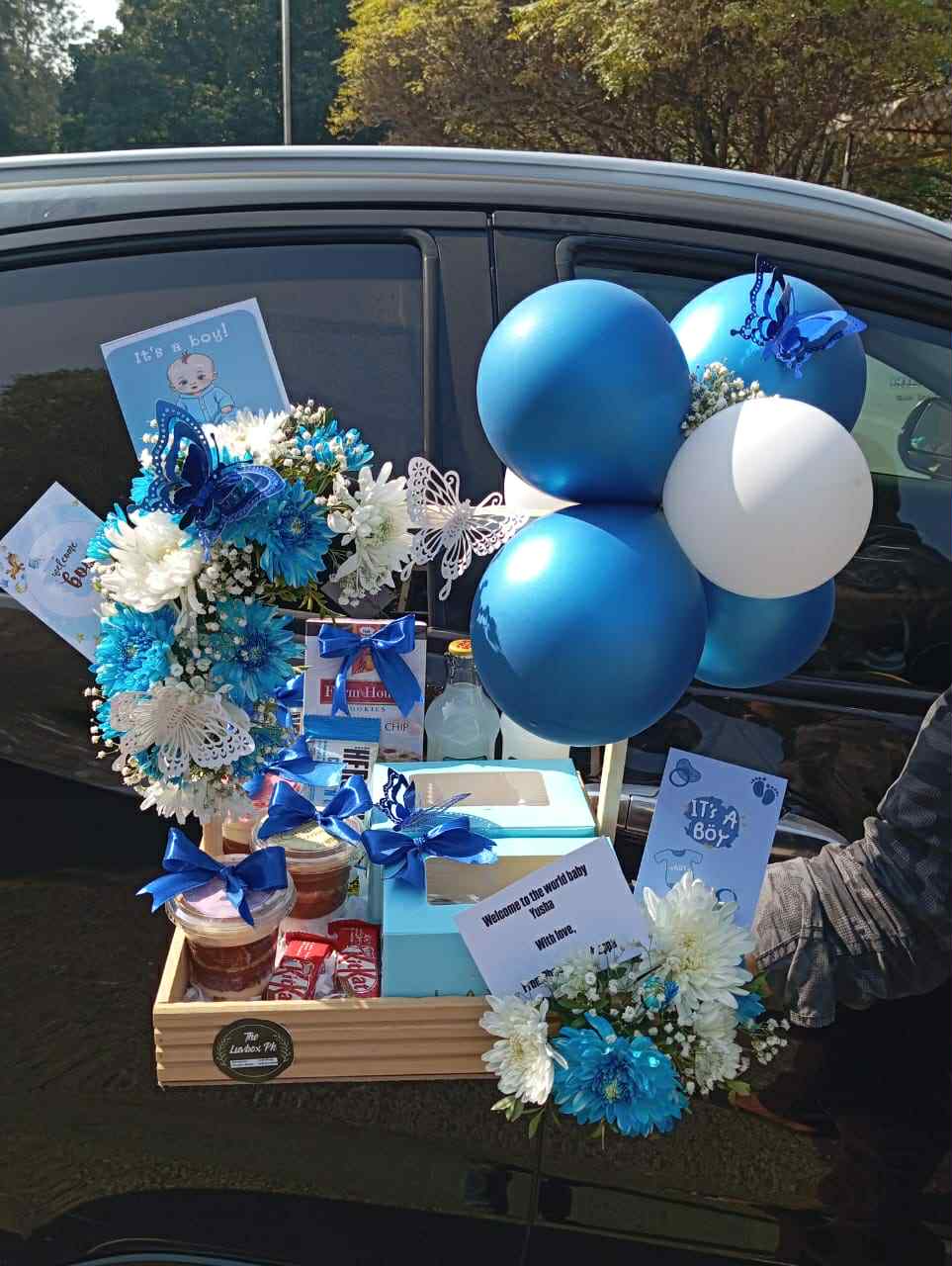 Wooden gift basket for baby boy celebration with blue flowers, cupcakes, brownies, chocolates, and drinks – perfect congratulations gift.